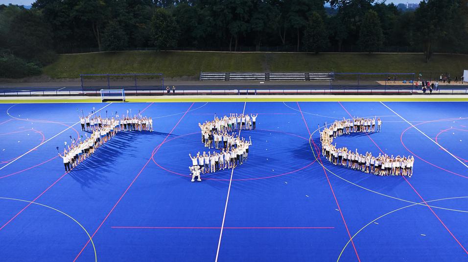 Ausschnitt einer Hockeyfeld-Inszenierung mit Menschen, die die Buchstaben "JSC" bilden, vor blauer Spielfeldfläche.