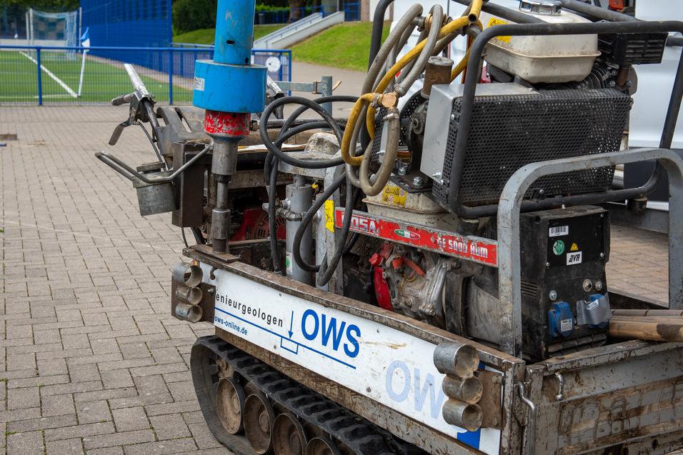Nahaufnahme einer Baustellenmaschine mit Schl&auml;uchen und Motor auf einem gepflasterten Bereich neben einem Fu&szlig;ballfeld.
