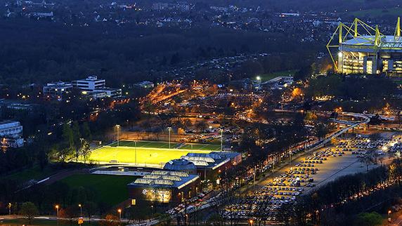 Luftaufnahme des Signal Iduna Parks mit beleuchtetem Stadion und umliegenden Parkplätzen bei Dämmerung.