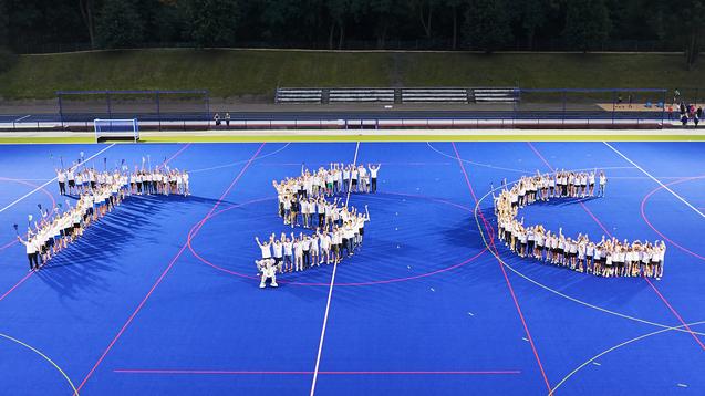 Ausschnitt einer Hockeyfeld-Inszenierung mit Menschen, die die Buchstaben "JSC" bilden, vor blauer Spielfeldfl&auml;che.
