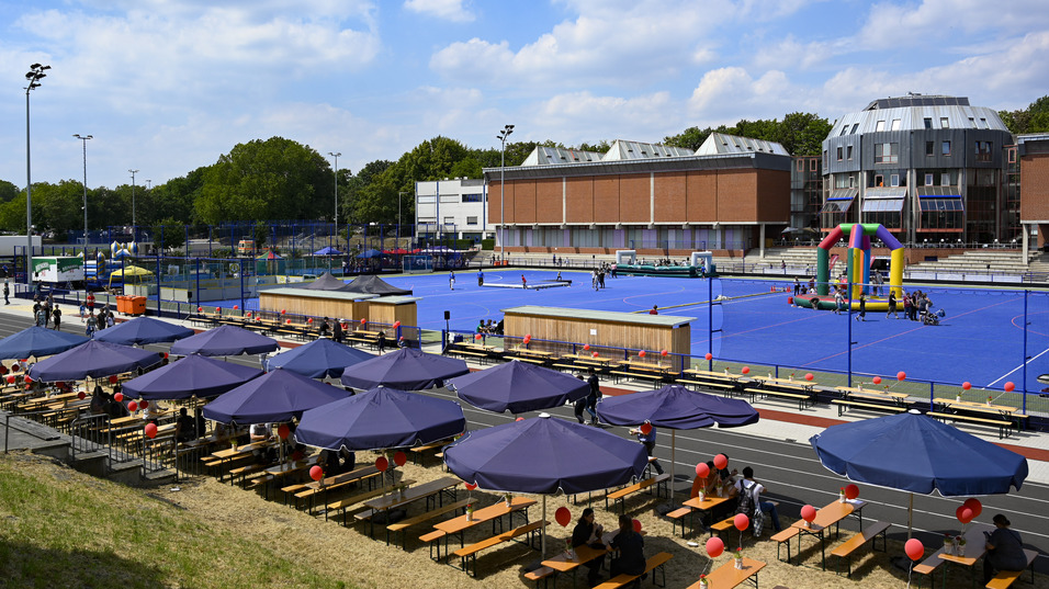 Blick auf ein Sportgel&auml;nde mit blauer Spielfl&auml;che, Sonnenschirmen und Sitzb&auml;nken, umgeben von gr&uuml;nen B&auml;umen.