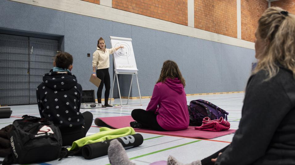 Eine Trainerin steht vor einer Gruppe auf Yoga-Matten und erklärt eine Übung an einem Flipchart in einer Sporthalle.