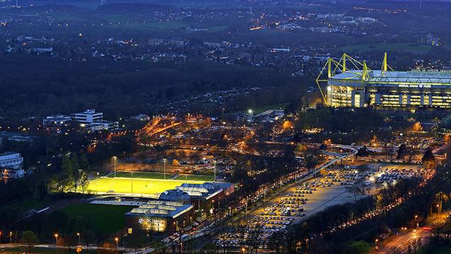 Luftaufnahme des Signal Iduna Parks mit beleuchtetem Stadion und umliegenden Parkpl&auml;tzen bei D&auml;mmerung.