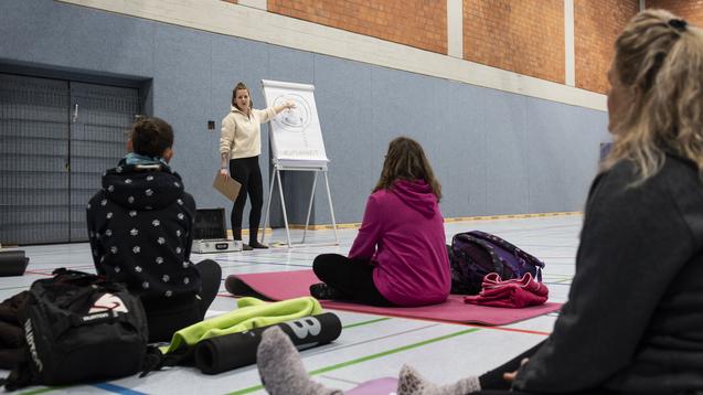 Eine Trainerin steht vor einer Gruppe auf Yoga-Matten und erkl&auml;rt eine &Uuml;bung an einem Flipchart in einer Sporthalle.