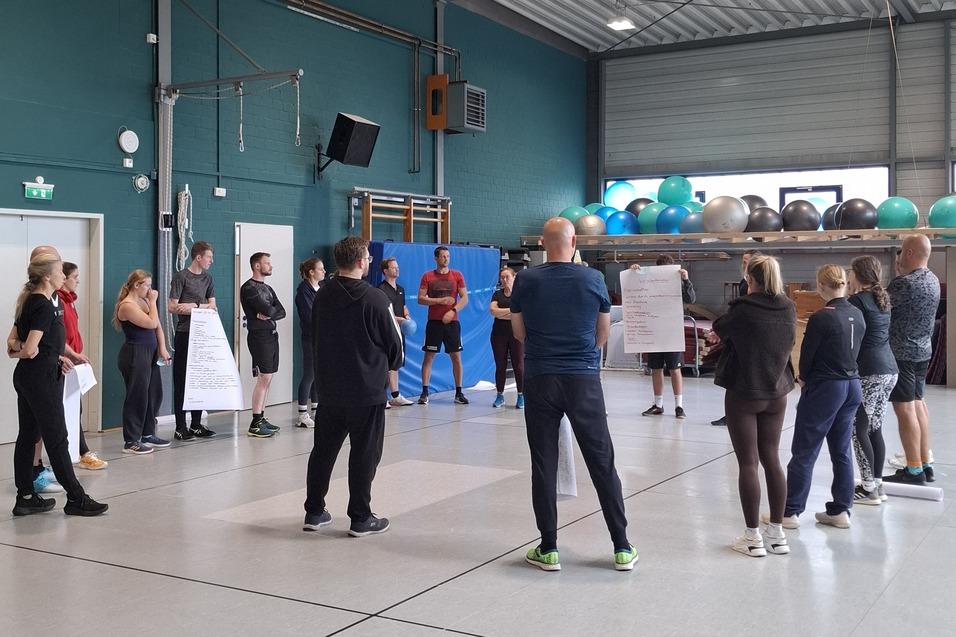 Gruppe von Menschen in Sportkleidung versammelt in einer gro&szlig;en Halle mit Fitnessger&auml;ten im Hintergrund.