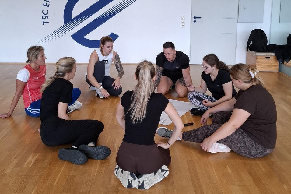Gruppe von T&auml;nzern sitzt im Kreis auf dem Boden in einem &Uuml;bungsraum mit dem Logo des TSC Eintracht Dortmund an der Wand.