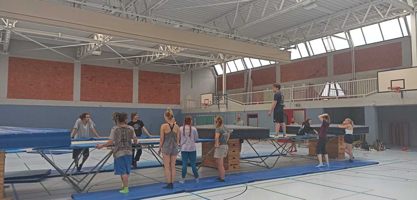 Gruppe von Sportlern, die an Trampolinen in einer hellen Sporthalle trainieren, mit blauer Matte und Fensterlicht.