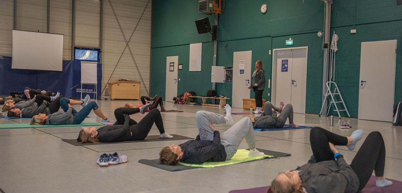 Gruppentraining in einer Halle, Teilnehmer liegen auf Matten und f&uuml;hren Dehn&uuml;bungen unter Anleitung aus.