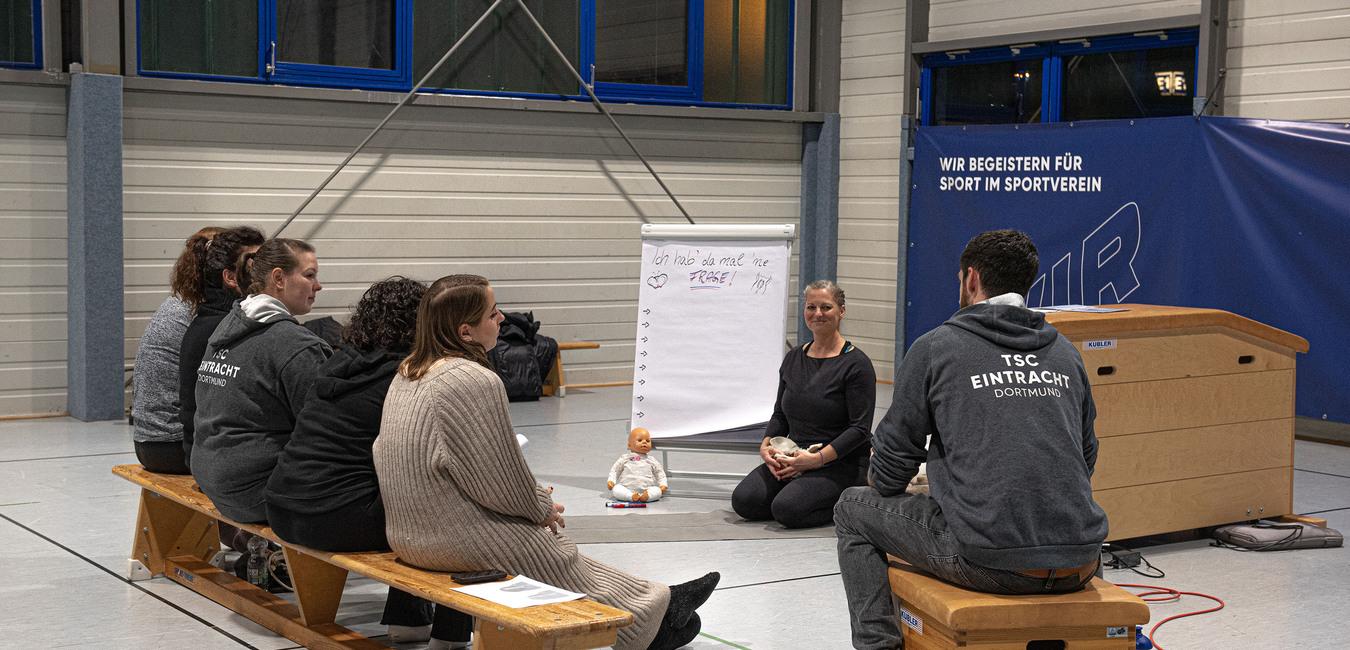 Gruppendiskussion in einer Turnhalle; acht Personen sitzen auf B&auml;nken, eine Frau pr&auml;sentiert an einem Flipchart.