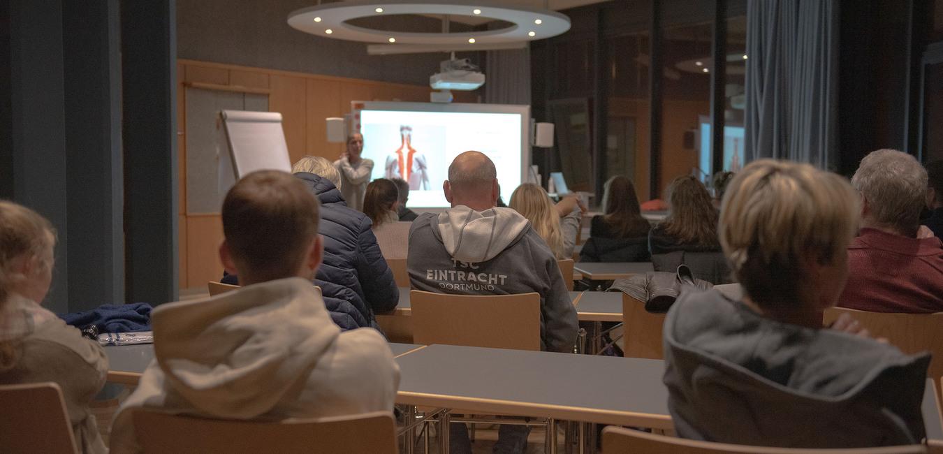Teilnehmende sitzen an Tischen in einem Seminarraum, w&auml;hrend eine Pr&auml;sentation an einer Wand gezeigt wird.