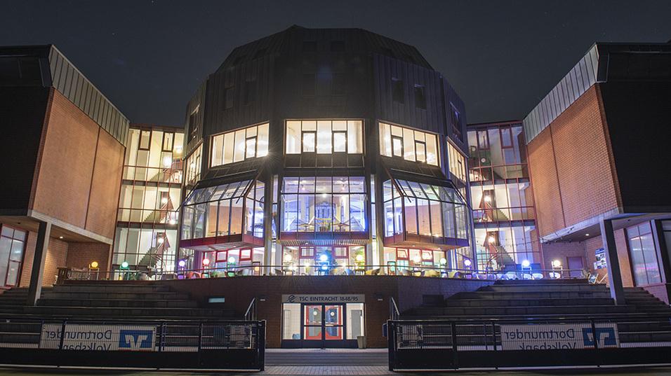 Beleuchtetes modernes Gebäude mit großen Fenstern und Terrassen, aufgenommen bei Nacht.