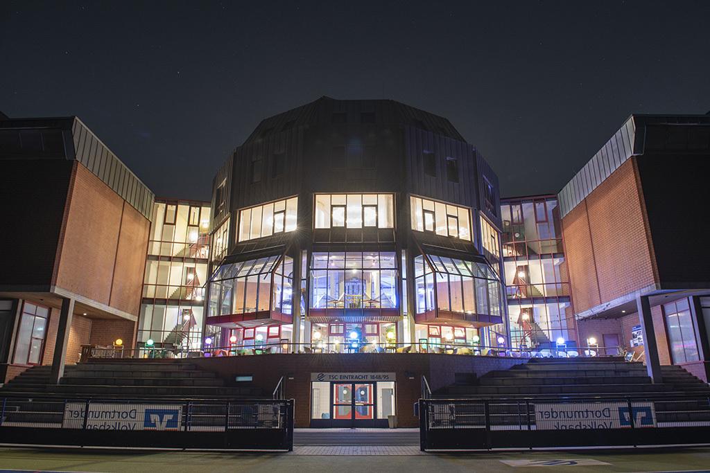Beleuchtetes modernes Gebäude mit großen Fenstern und Terrassen, aufgenommen bei Nacht.