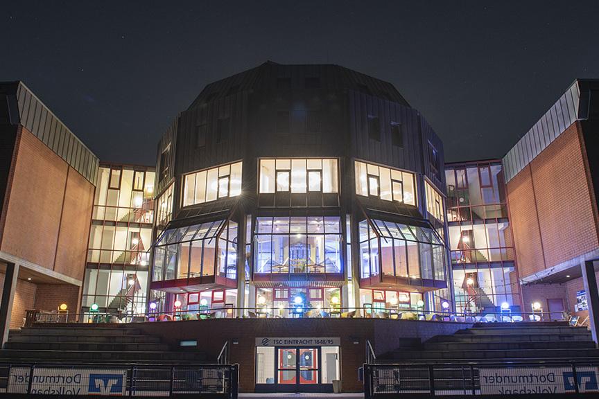 Beleuchtetes modernes Geb&auml;ude mit gro&szlig;en Fenstern und Terrassen, aufgenommen bei Nacht.