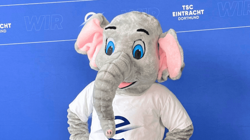 Maskottchen eines Elefanten in Sportkleidung steht vor einem blauen Hintergrund mit dem Logo des TSC Eintracht Dortmund.