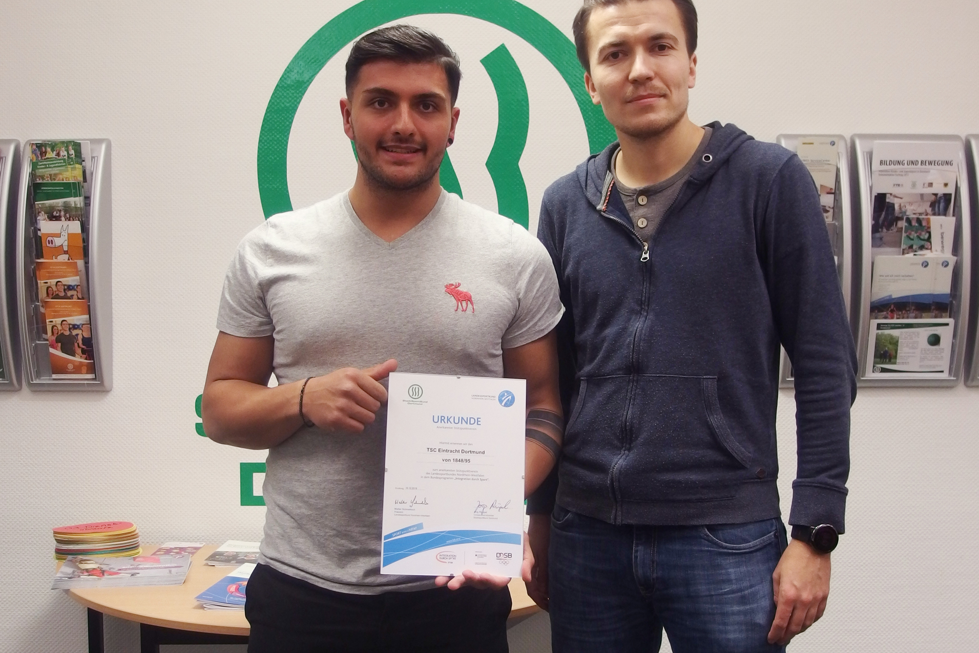 Zwei M&auml;nner stehen in einem B&uuml;ro des StadtSportBunds Dortmund und halten eine Urkunde in der Hand.