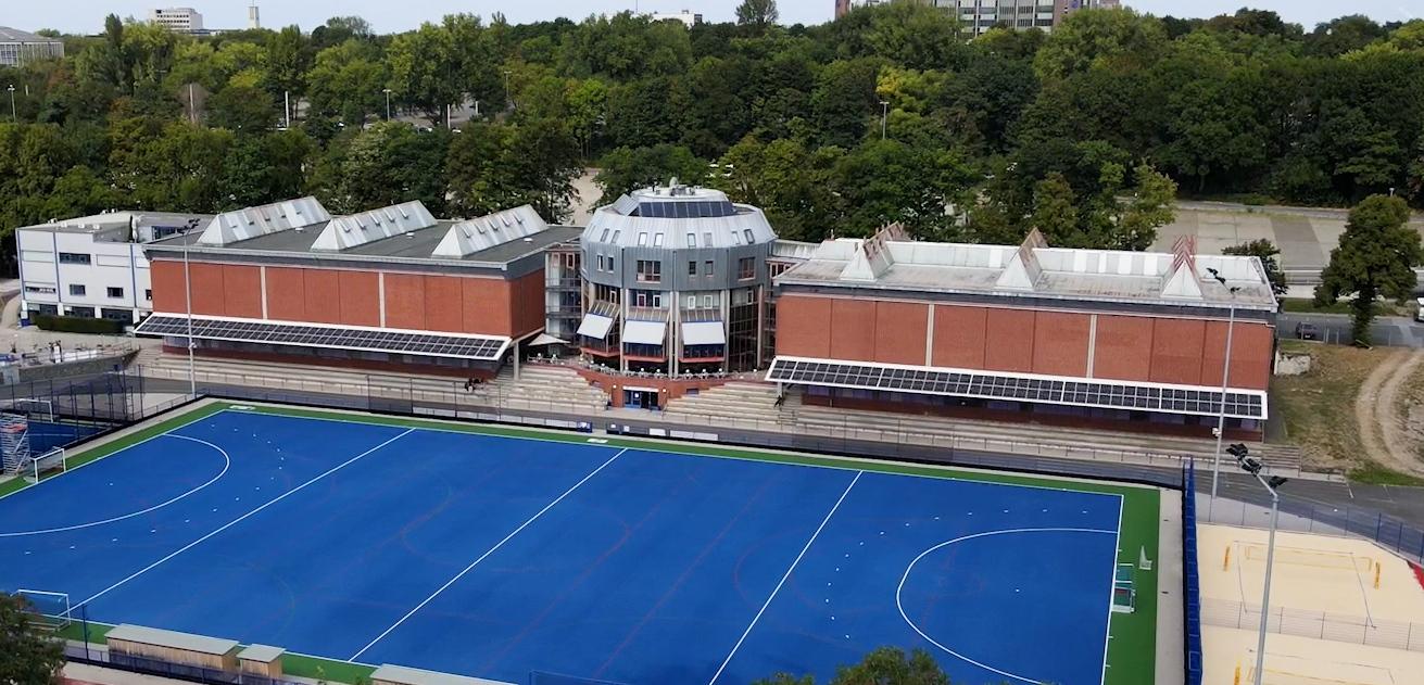 Luftaufnahme eines Sportgeländes mit blauer Hockeyplatz und modernen Gebäuden im Hintergrund, umgeben von Bäumen.