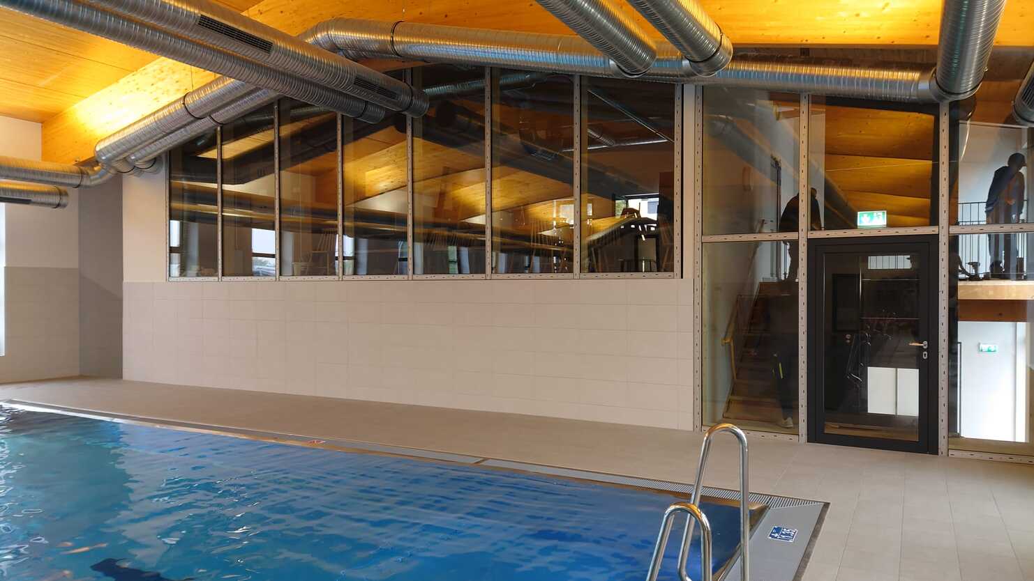 Innenansicht eines Schwimmbads mit blauer Wasseroberfl&auml;che und gro&szlig;en Fenstern, die Tageslicht einlassen.