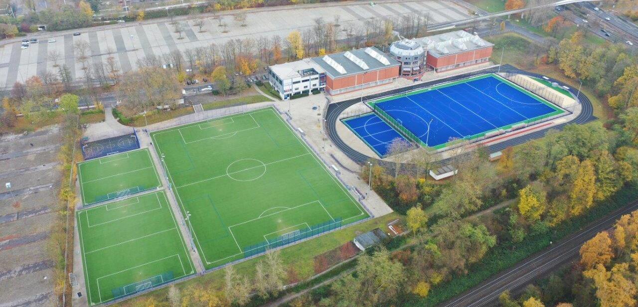 Luftaufnahme eines Sportgel&auml;ndes mit zwei Fu&szlig;ballfeldern und einem Hockeyplatz neben einem modernen Geb&auml;ude.