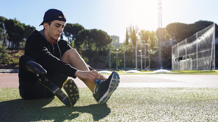 Junger Athlet sitzt auf einem Sportplatz und bindet den Schnürsenkel seines Schuhs, daneben eine Prothese.