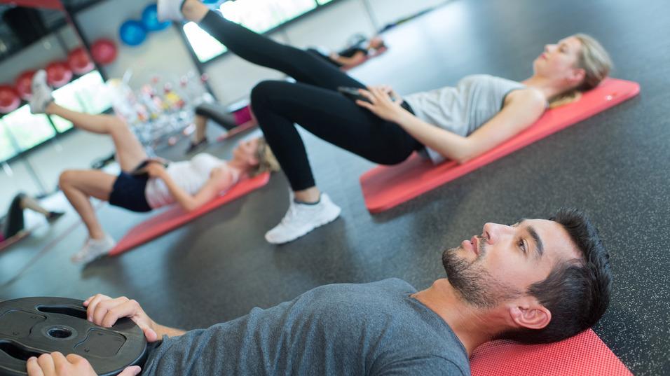 Gruppe von Menschen bei einem Workout, einige auf Fitnessmatten mit Gewichten, andere in Bauchlage.