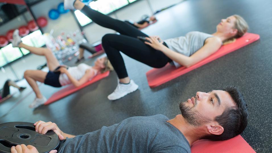 Gruppe von Menschen bei einem Workout, einige auf Fitnessmatten mit Gewichten, andere in Bauchlage.