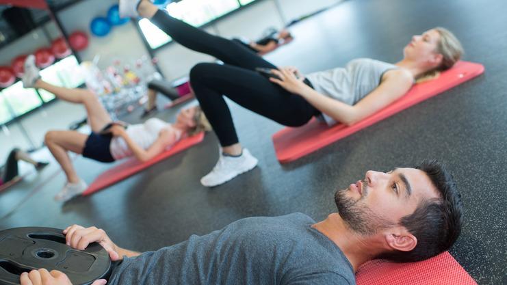 Gruppe von Menschen bei einem Workout, einige auf Fitnessmatten mit Gewichten, andere in Bauchlage.