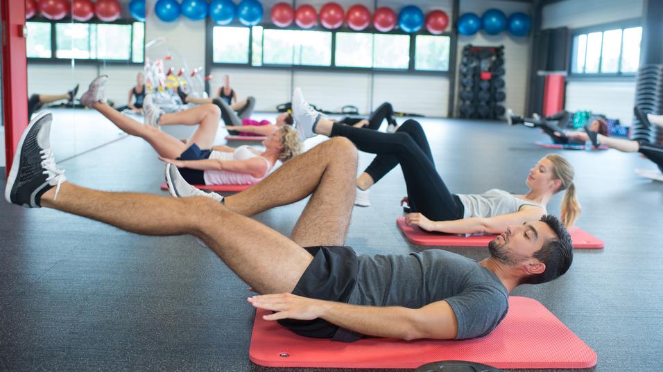 Gruppe von Personen macht Übungen auf Fitnessmatten in einem modernen Gym mit Fitnessgeräten im Hintergrund.