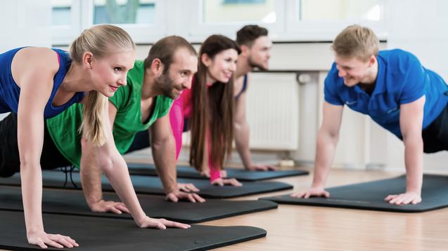 Fünf Personen in Sportbekleidung machen Liegestütze auf Matten in einem hellen Fitnessraum.