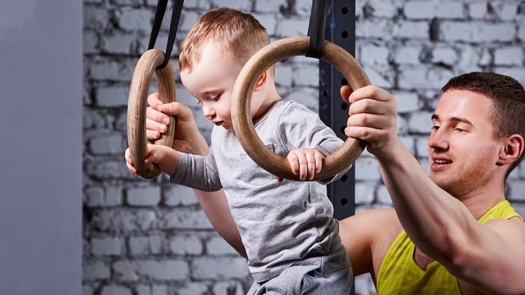 Ein Baby schwingt an Holzturnringen, während ein Erwachsener unterstützend daneben steht. Hintergrund mit Ziegelmauer.