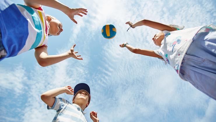 Drei Kinder werfen einen bunten Volleyball in den Himmel vor einem blauen Himmel mit einigen Wolken.