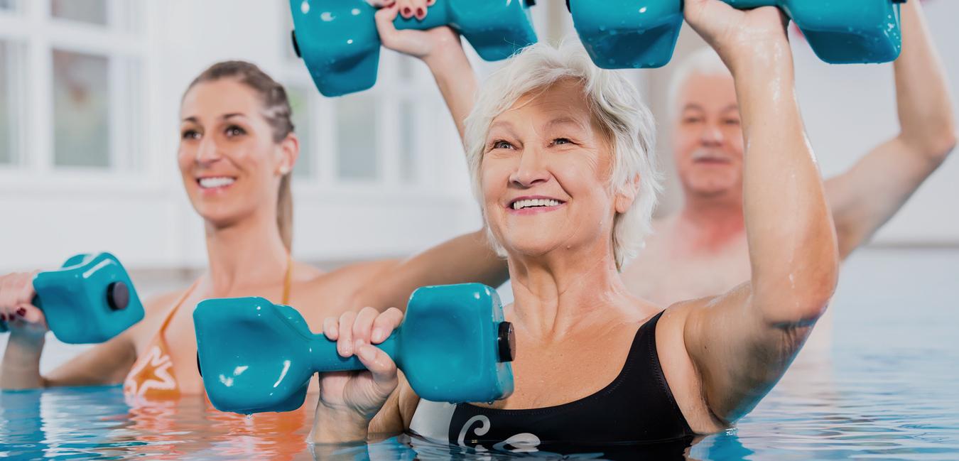 Drei Personen im Wasser heben blaue Hanteln, lächeln und üben Aquafitness in einem hellen Schwimmbad.