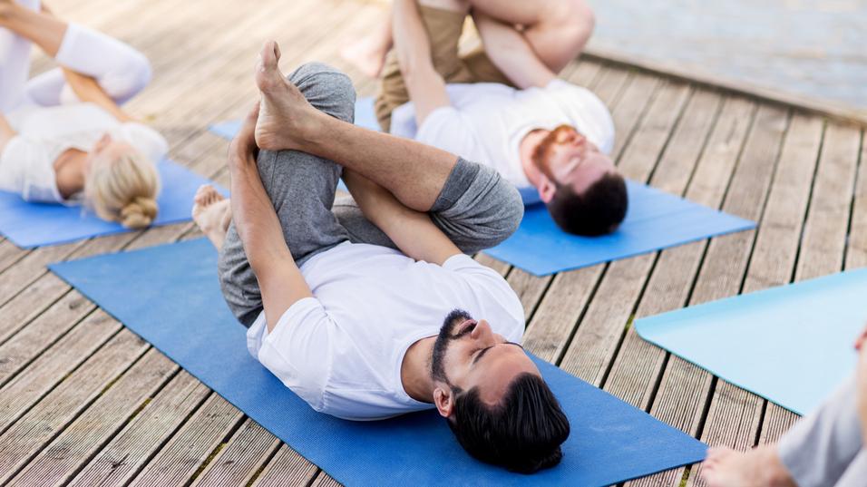 Gruppe von Menschen in Sportkleidung macht Yoga auf blauen Matten auf einem Holzsteig neben einem Gewässer.